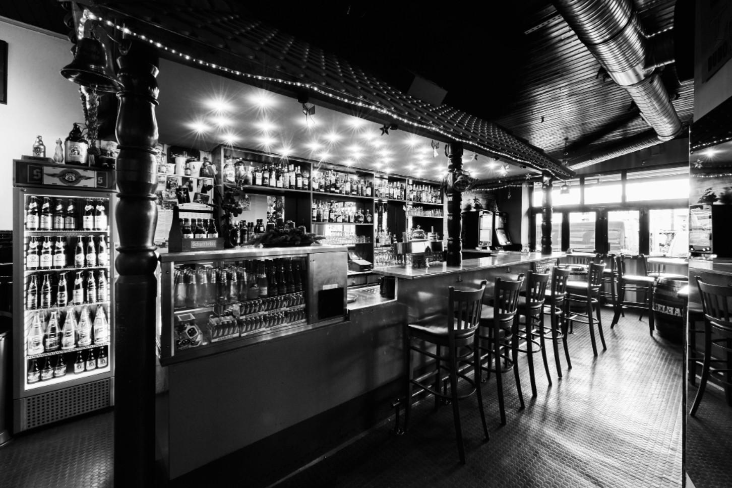 Eine Bar mit zahlreichen Flaschen und Gläsern auf Regalen sowie Hockern vor der Theke. Die Beleuchtung ist punktuell und schafft helle Lichteffekte.