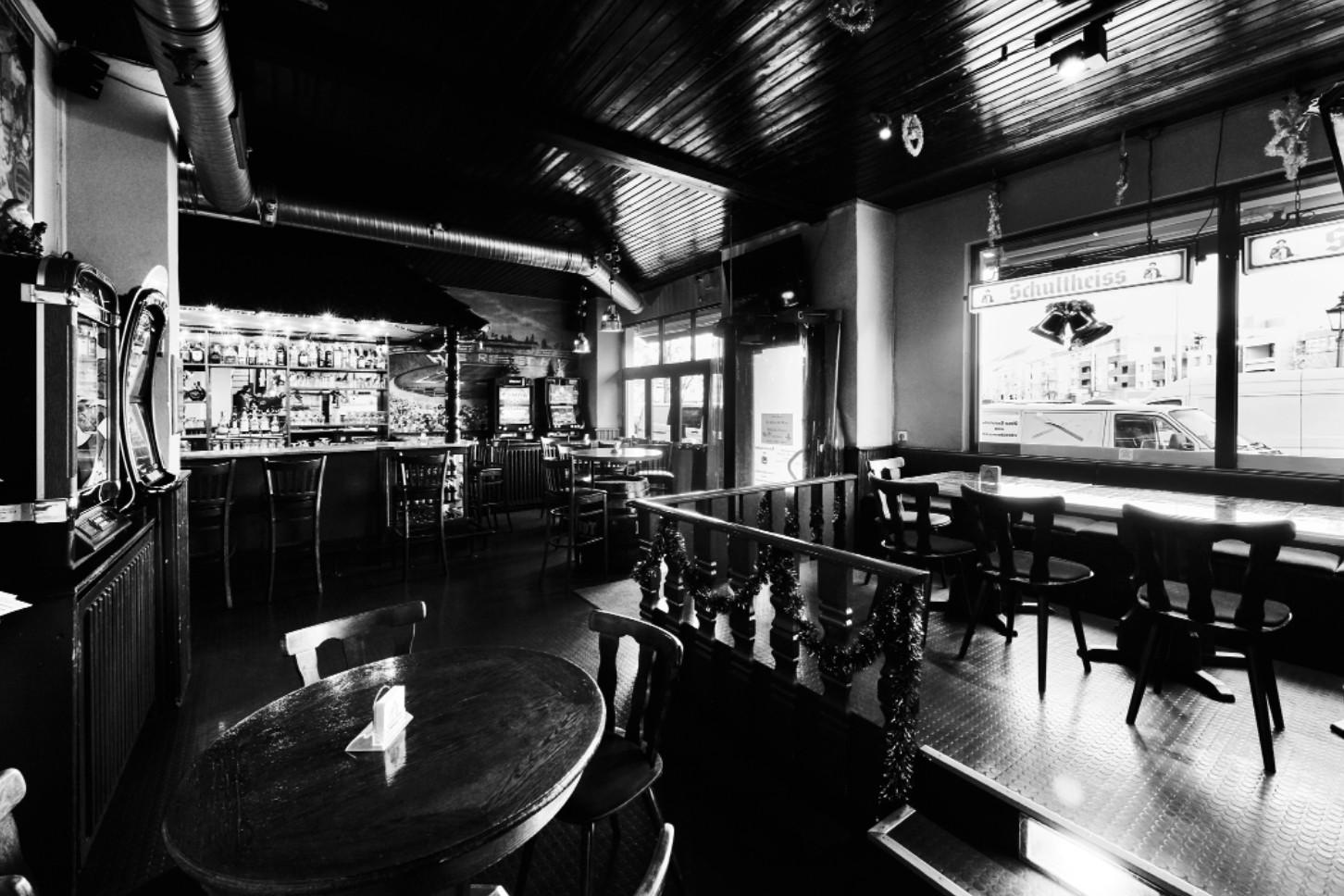 Innenansicht einer Bar mit Holztischen, einer Theke und großen Fenstern zur Straße.