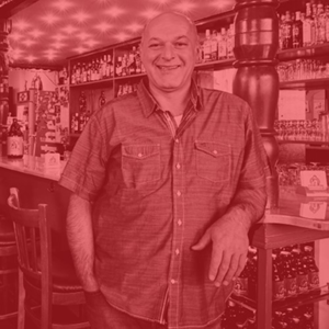 Ein lächelnder Mann steht vor einer Bar mit vielen Flaschen im Hintergrund.