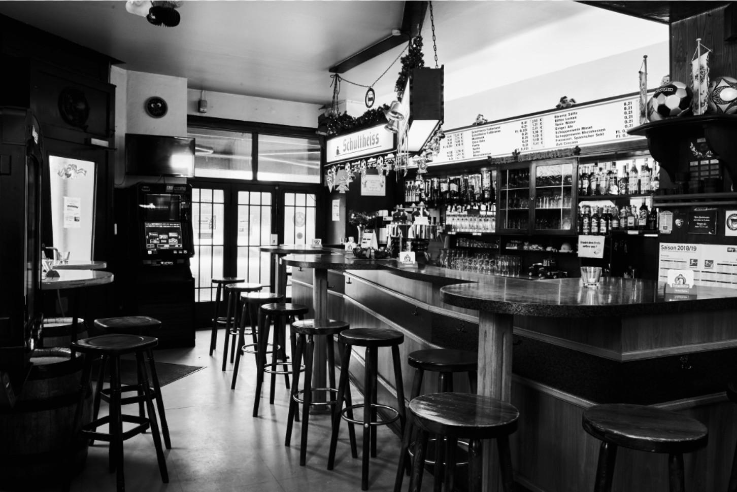 Ein schwarz-weißes Foto zeigt das Innere einer Bar mit Theke, Hockern und Getränkeregalen.