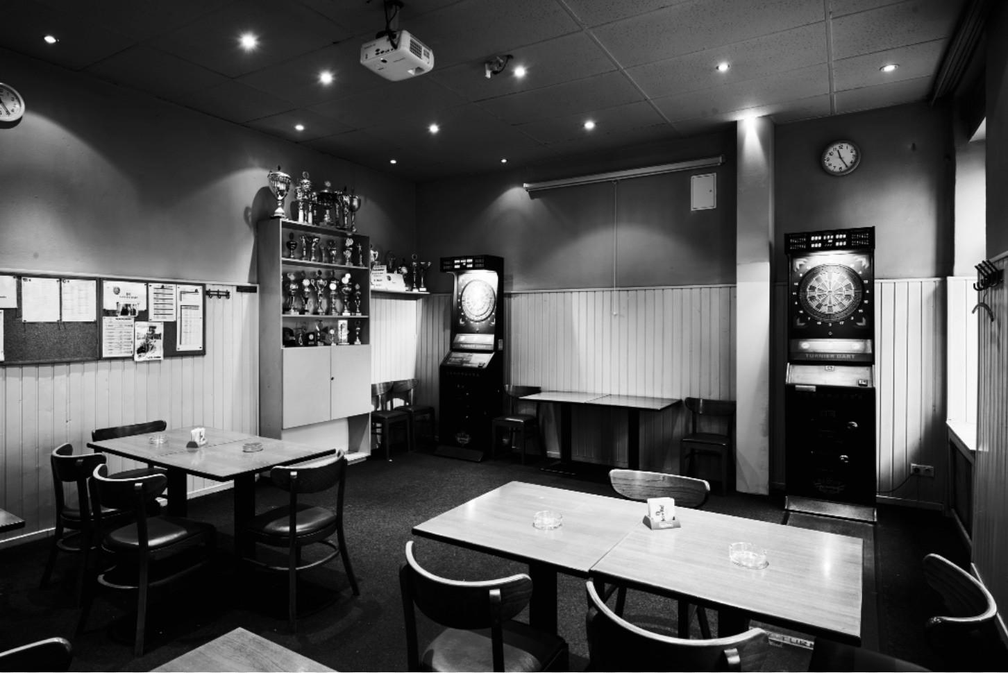 Ein leeres Café mit Holztischen, schwarzen Stühlen und einem Dartautomaten. An der Wand hängen Trophäen und Aushänge.