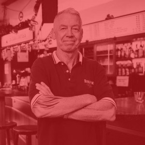 Ein älterer Mann steht mit verschränkten Armen an einer Bar.