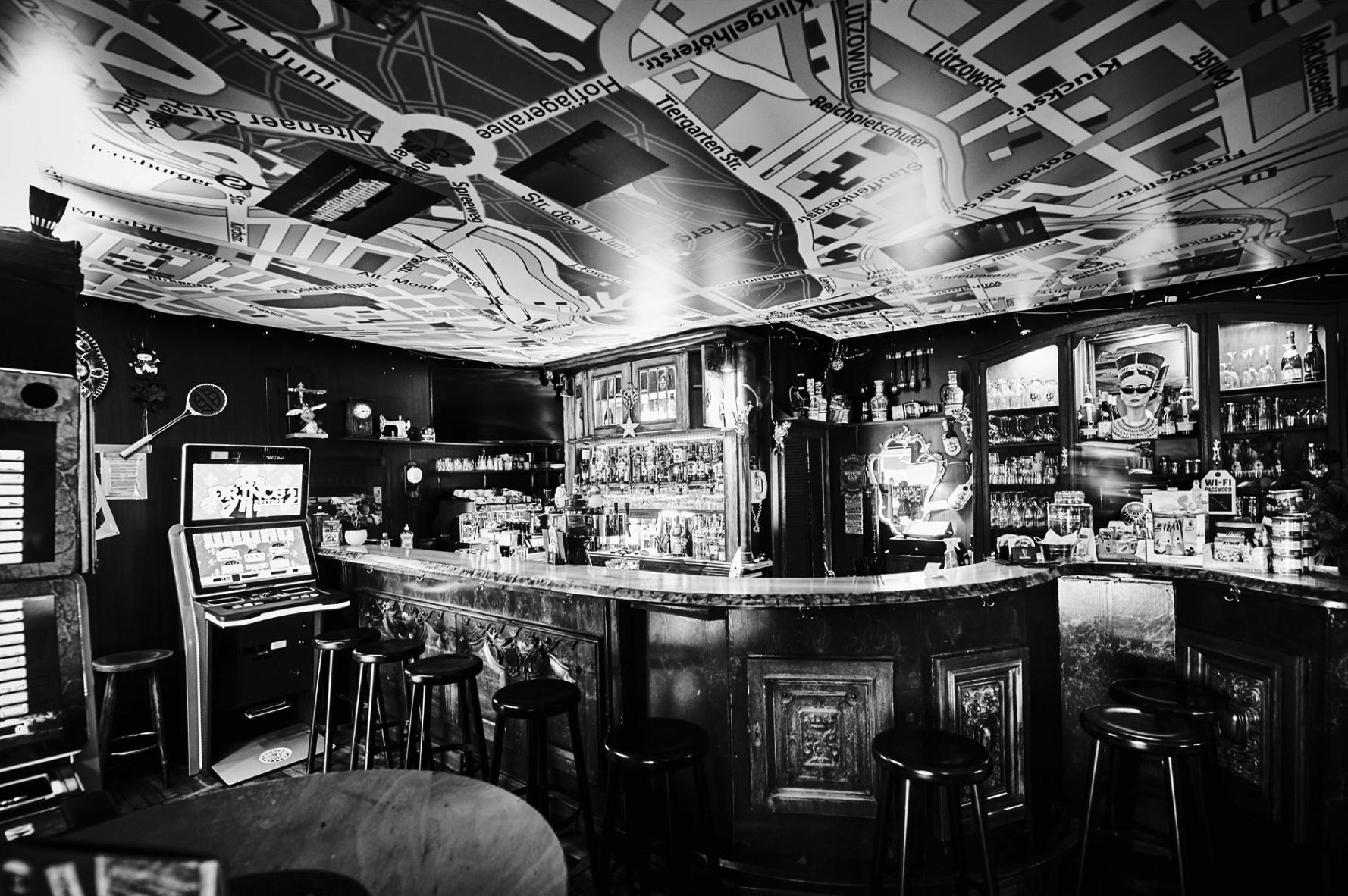 Eine Bar mit einem detaillierten, dunklen Interieur und Postern an der Decke. An der Bar stehen Hocker und ein Spielautomat ist sichtbar.