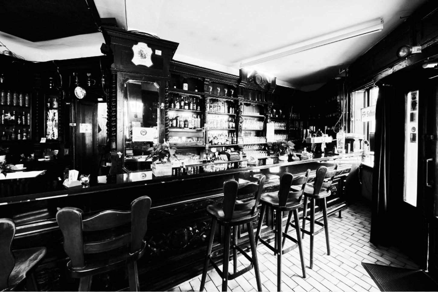 Eine klassische Bar mit Holztheke und Hochstühlen ist abgebildet. Im Hintergrund sind Regale voller Flaschen zu sehen.