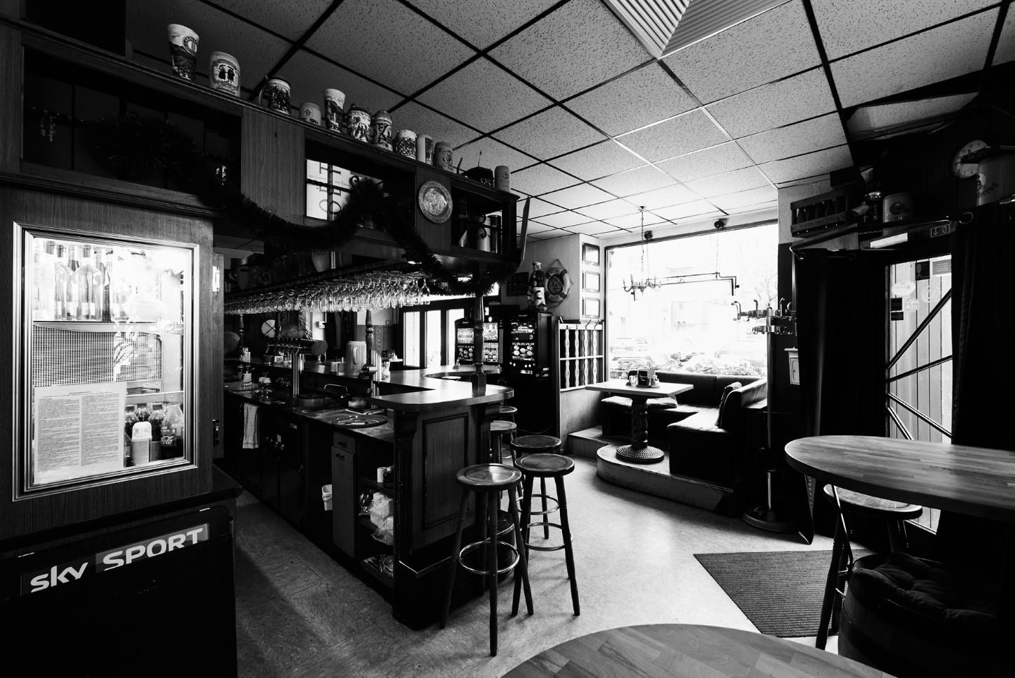 Eine schwarz-weiße Innenaufnahme zeigt eine gemütliche Bar mit Stühlen, Tischen und einer Theke. Verschiedene Gläser und Tassen sind sichtbar.