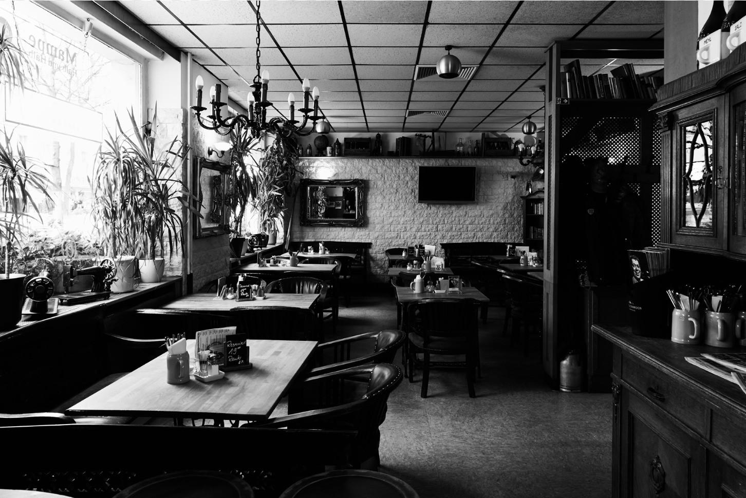 Die Szene zeigt ein Café mit vielen Tischen, Stühlen und Deckenlampen. Pflanzen und Gemälde dekorieren den Raum.