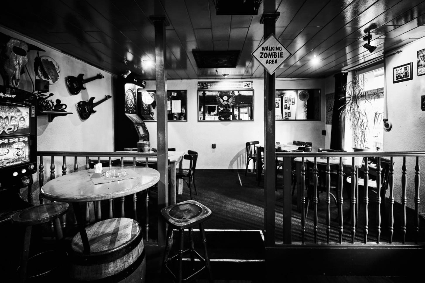 Ein schwarz-weißes Foto zeigt das Innere einer Bar mit Tischen, Stühlen und Gitarren an den Wänden.