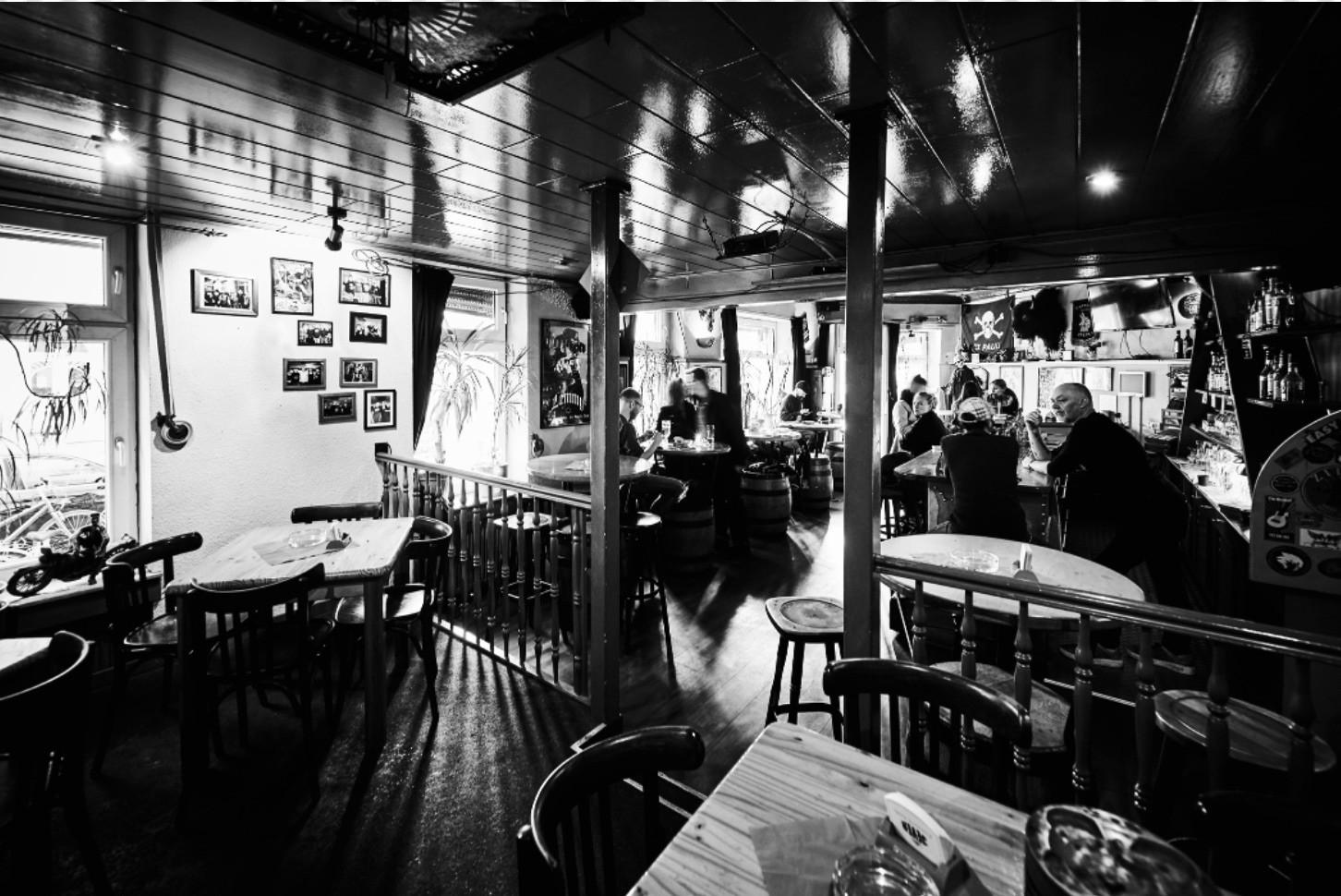 Innenansicht einer belebten Bar mit Gästen an Tischen und der Theke, Wände mit Bildern dekoriert.
