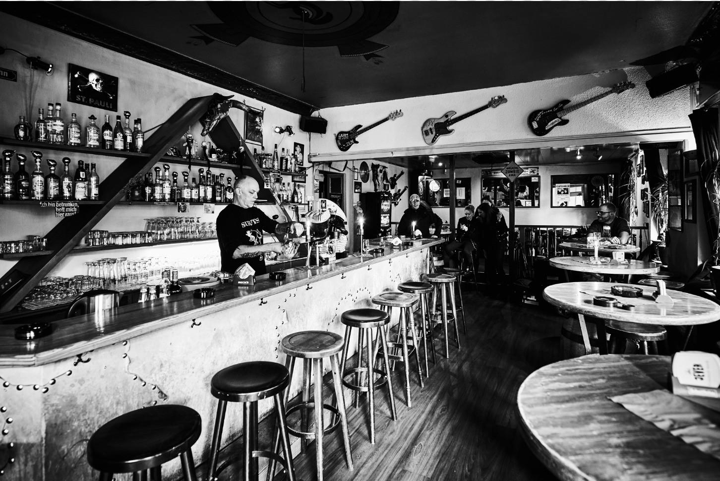 Eine Bar mit mehreren Gästen und einem Barkeeper, der ein Getränk zubereitet. An der Wand hängen Gitarren.