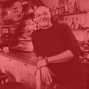 Ein lächelnder Mann lehnt an einer Bar mit vielen Flaschen im Hintergrund.