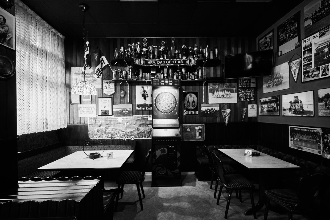 Ein leerer Kneipenraum mit Trophäen und historischen Fotos an den Wänden. In der Mitte stehen Tische und Stühle.