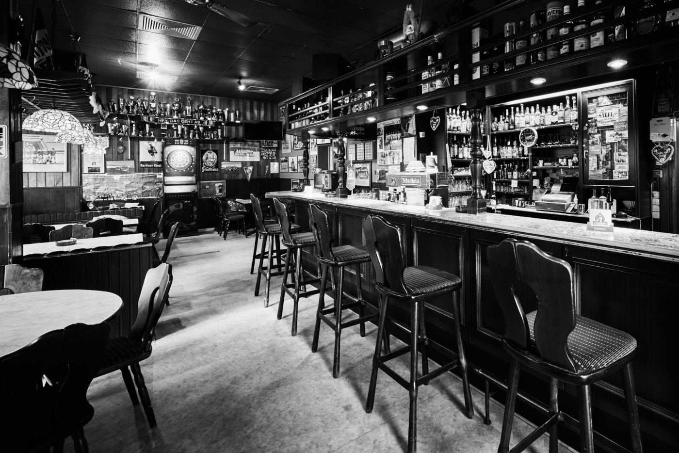 Mehrere Barhocker stehen vor einer Theke in einem traditionellen Pub. An der Wand hängen zahlreiche Dekorationsgegenstände.