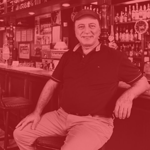 Ein älterer Mann sitzt lächelnd an einer Bar. Er trägt eine Mütze und ein Poloshirt.